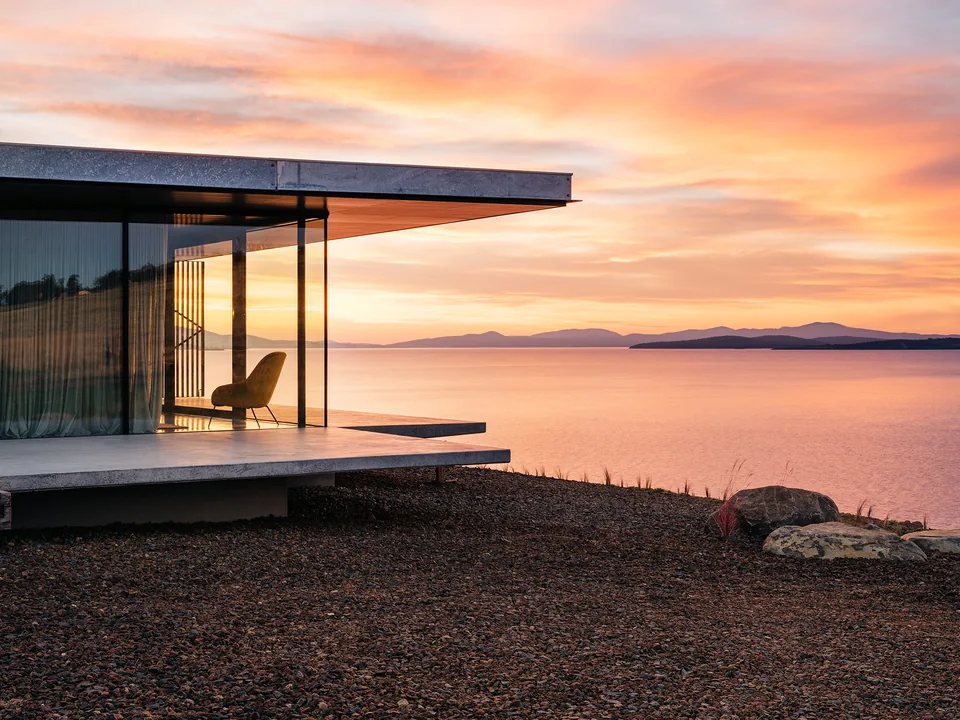 A glass-walled pavilion home on a concrete platform at the water's edge, one chair inside, under a peach sunset over distant hills