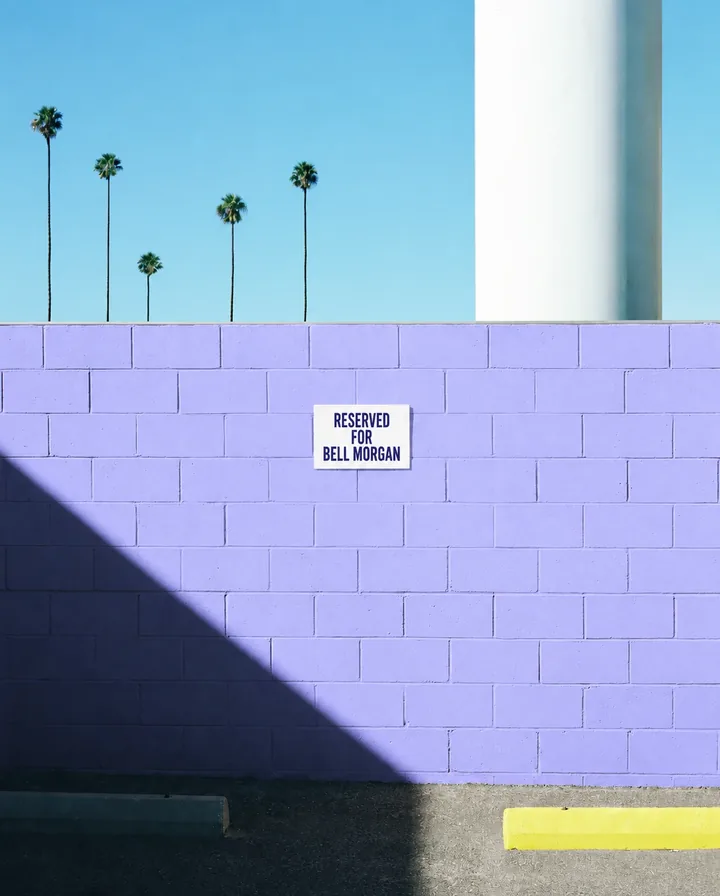 A periwinkle block wall under palm trees with a small sign reading Reserved for Bell Morgan